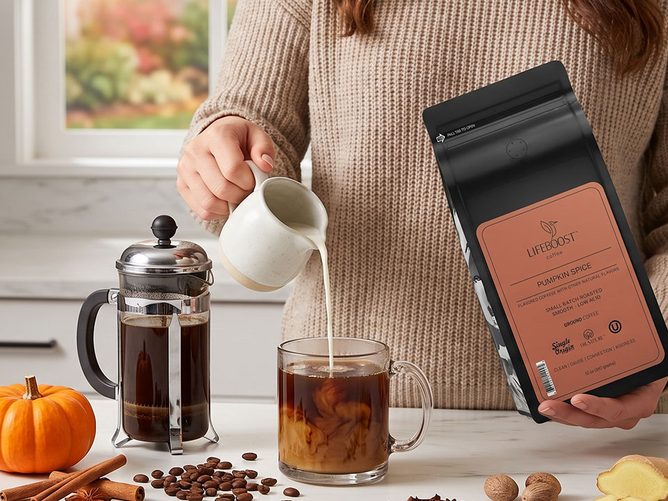 Pouring cream into a glass mug of Lifeboost Pumpkin Spice coffee beside a French press and the Pumpkin Spice bag